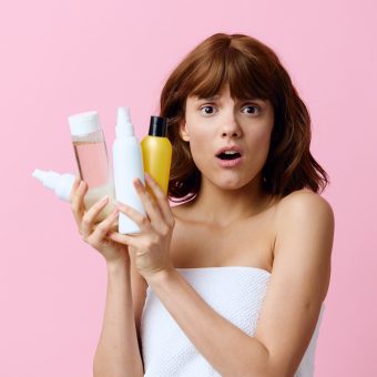horizontal photo on a pink background of a shocked, surprised woman, holding various care products in her hands and looking at the camera with her mouth wide open